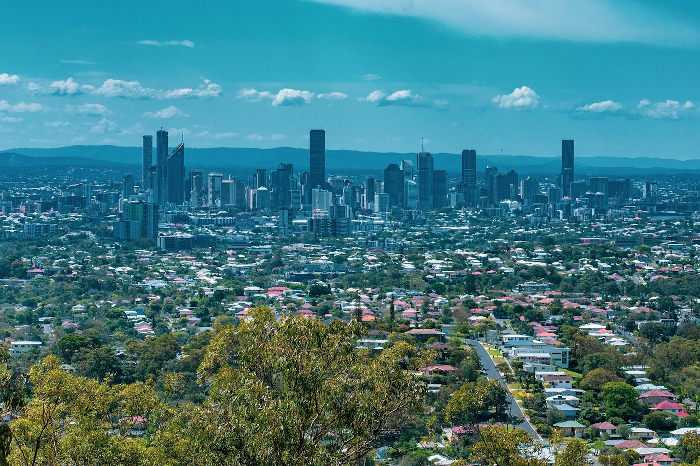 Panoramic view of Brisbane, Australia, showing suburban homes in the foreground and the city skyline with high-rise buildings in the background under a clear blue sky.