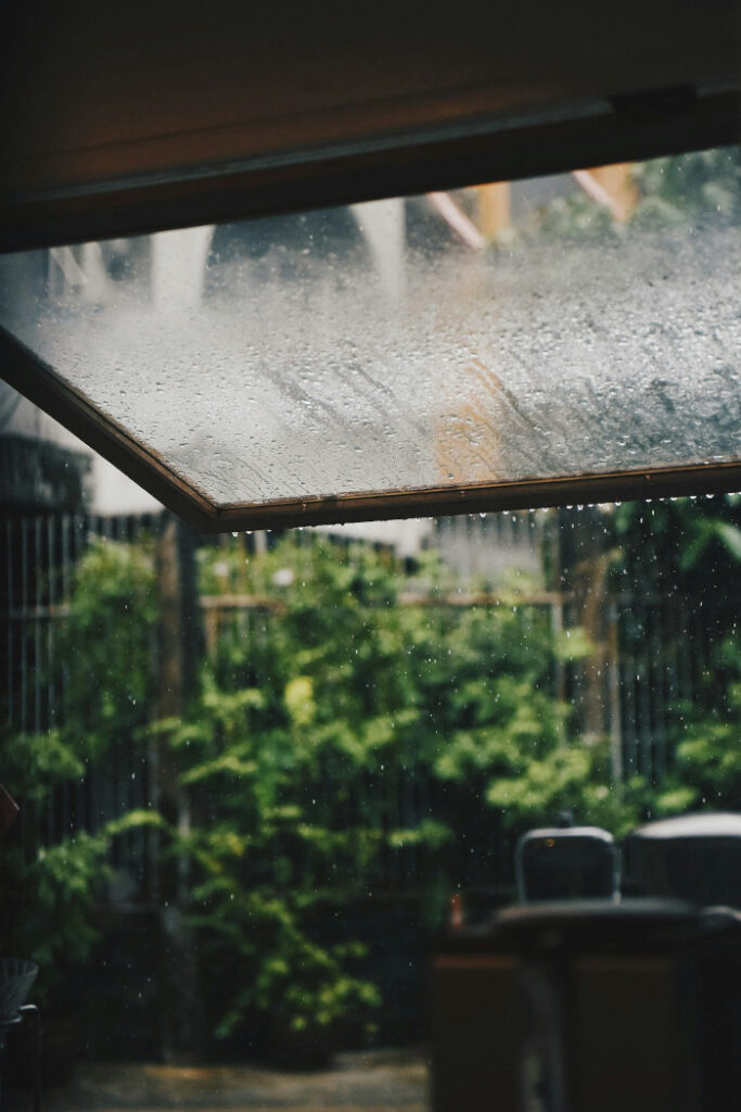View through a partially open window on a rainy day, with water droplets on the glass and a blurred garden in the background.
