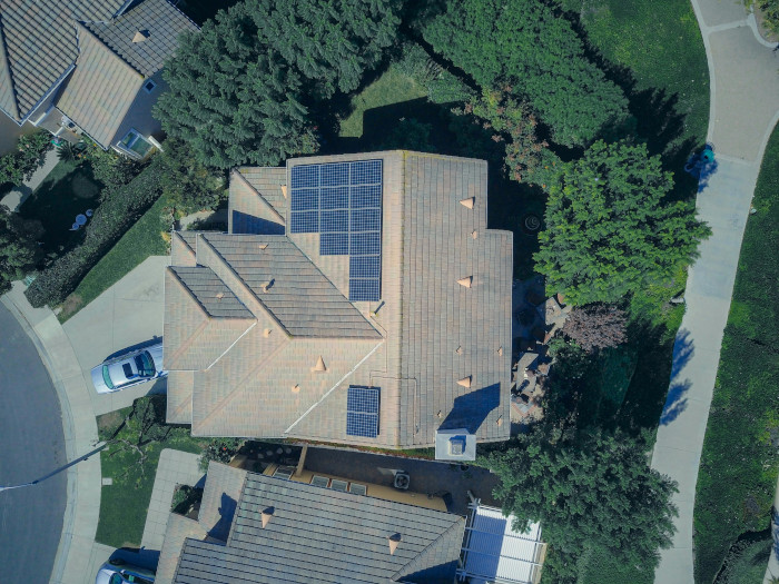 Aerial view of a residential house with multiple solar panels installed on the roof, surrounded by trees and neighboring homes.