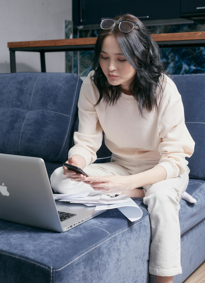 Woman sitting on a blue couch using a smartphone while reviewing bills and documents with a laptop in front of her.
